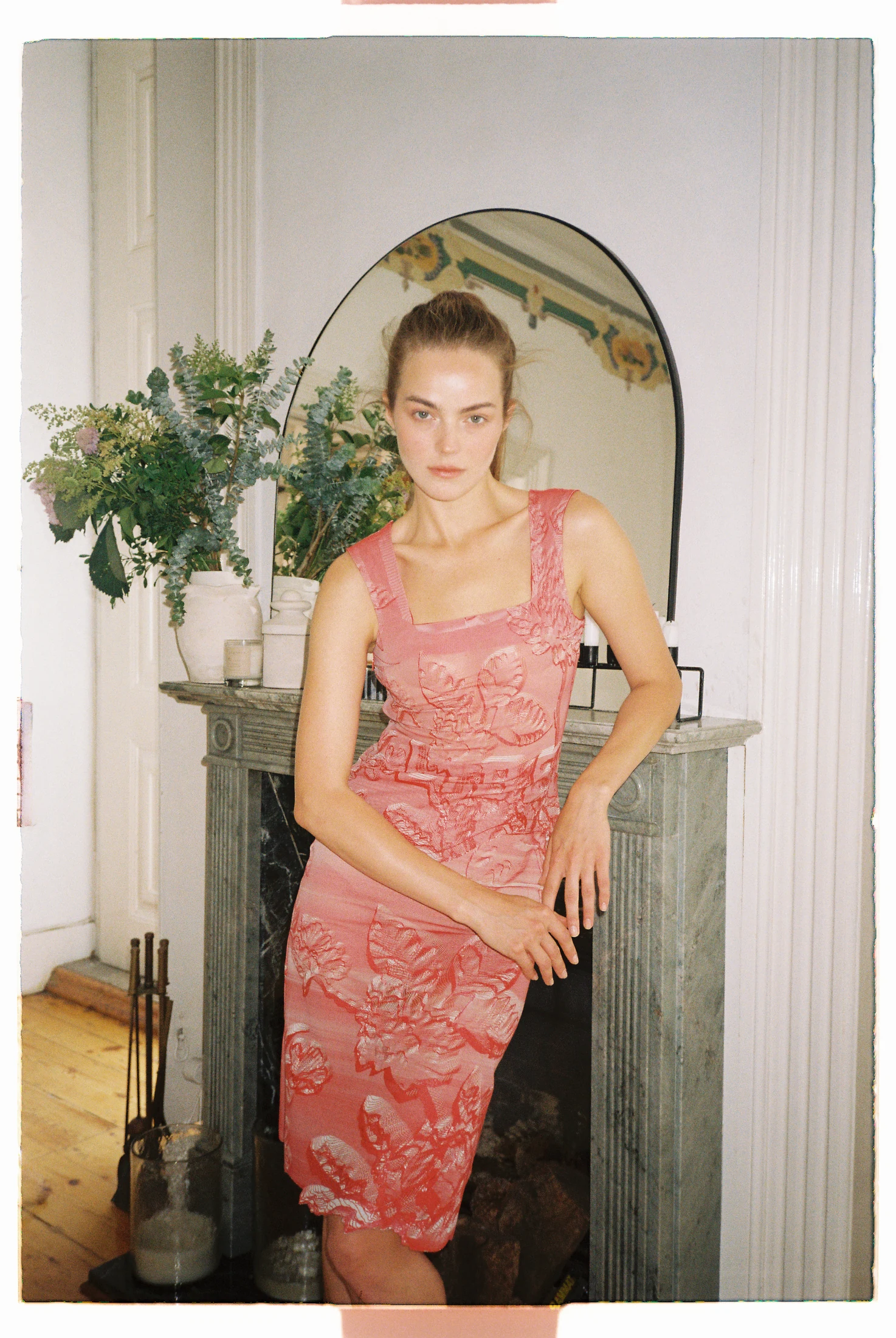 A model wearing a pink floral 3D-printed dress beside a fireplace and mirror